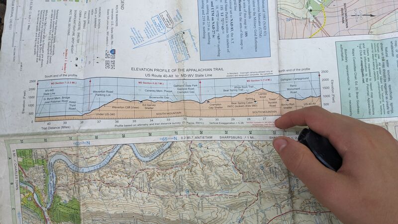The image shows a person's hand holding a magnifying glass over a map. The map appears to be a topographical map, possibly for hiking or navigation, with elevation profiles and geographical details. The person seems to be examining the map closely, likely for route planning or orientation purposes.